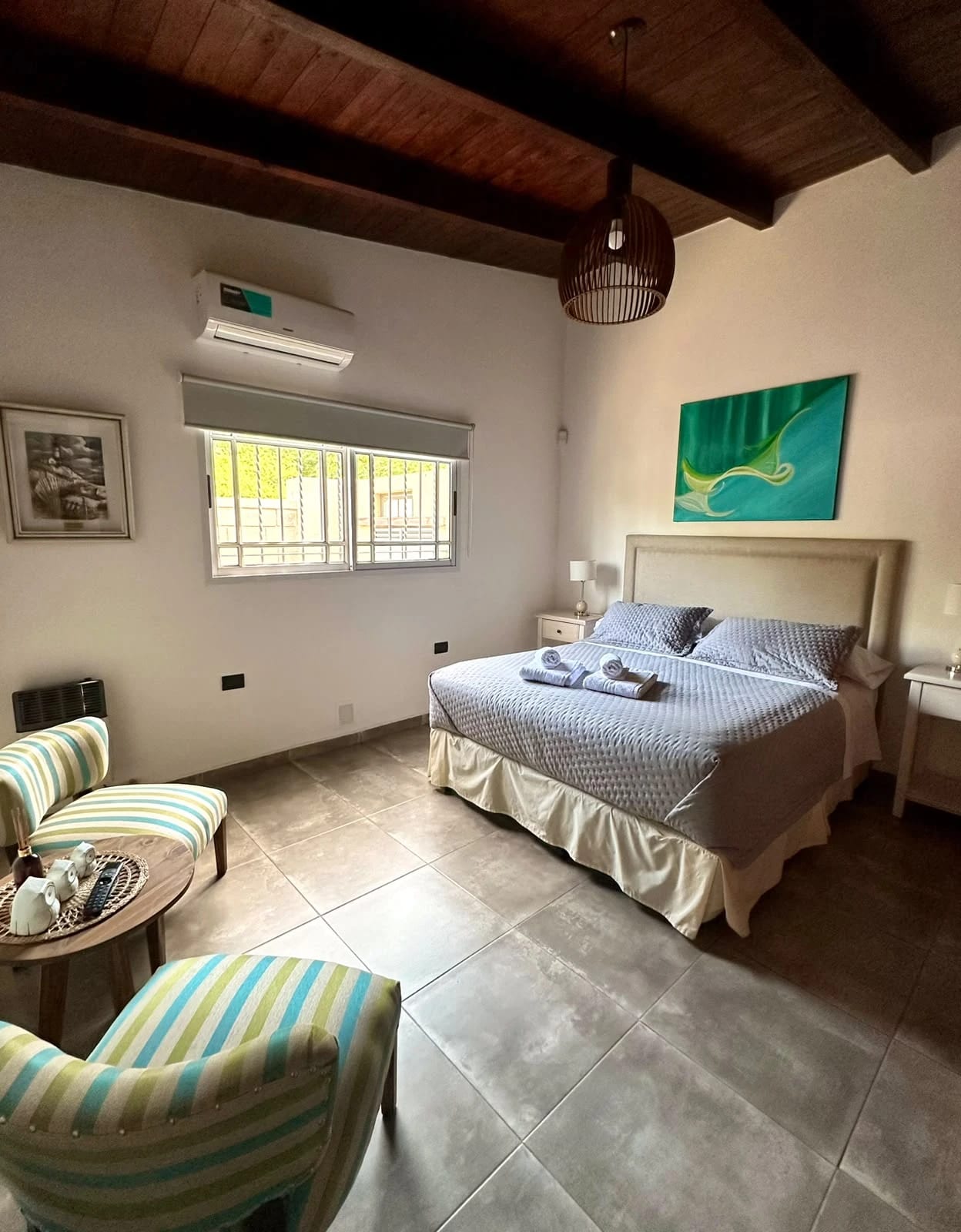 Habitación matrimonial con cama tendida, toallas dobladas, sillones y techo de madera